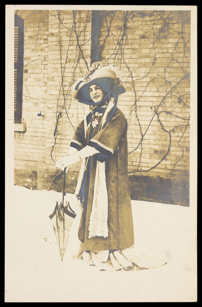 A man in drag, wearing a large bonnet, leans on a parasol whilst standing in front of a brick building. Photographic…