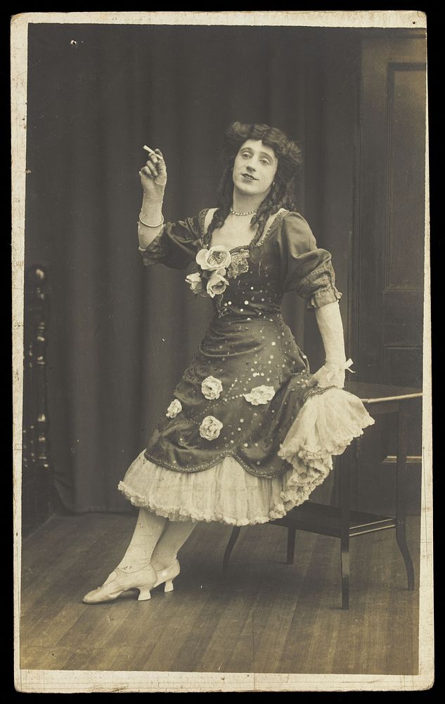A performer in drag, wearing a large flowery dress, poses with a cigarette on stage. Photographic postcard, ca. 1908-1910.