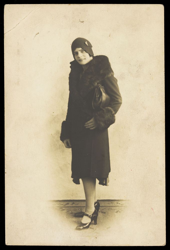 A person poses wearing a long coat with fur trim and a hat with a brooch.