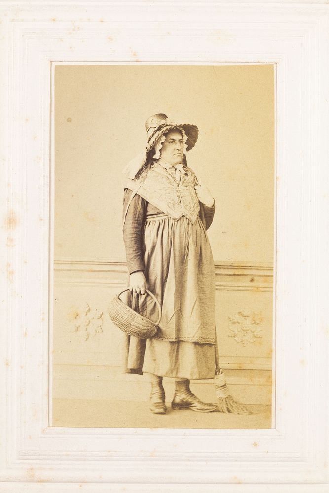 A man in drag poses with a bonnet, basket and broom. Photograph, 189-.