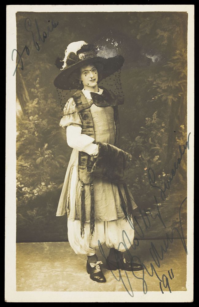 A man in drag wearing a very large hat with a veil, stands in front of a painted backdrop, with dense foliage. Photographic…