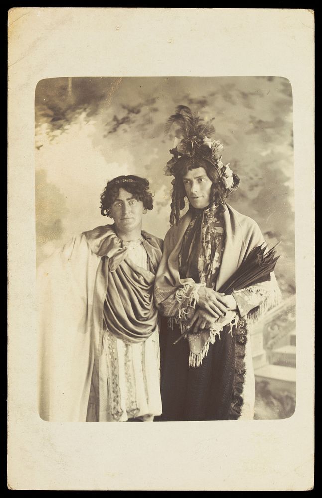 Two men in drag, one wearing a detailed feathered head garment, pose for a portrait. Photographic postcard by F. Wood, 190-.