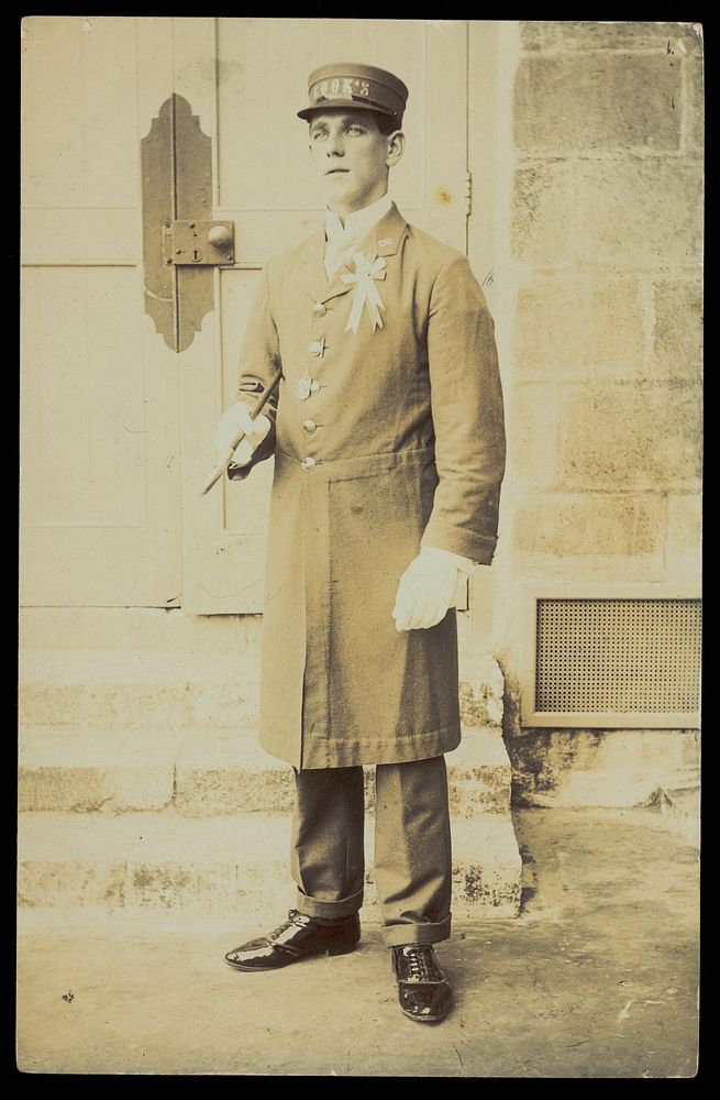 A man dressed wearing a long coat and a hat bearing the word "Cook", stands outside a building. Photographic postcard, 191-.