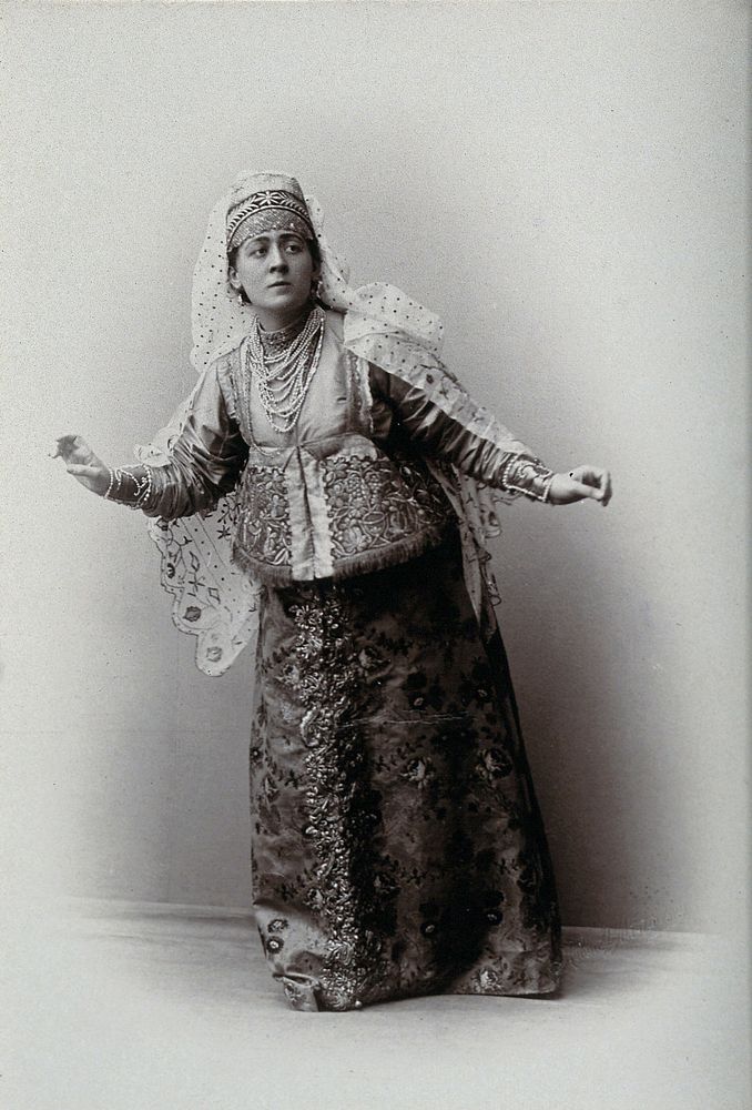 A woman posing in a photographic studio, wearing an embriodered dress.