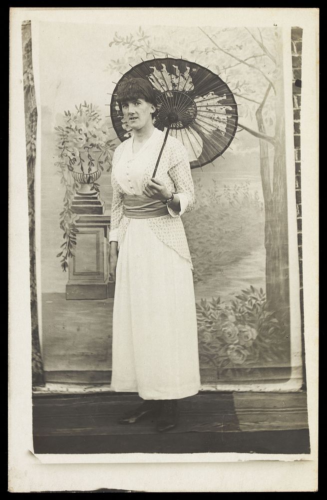 An amateur actor in drag poses with an open parasol, in front of painted scenery. Photographic postcard, 191-.