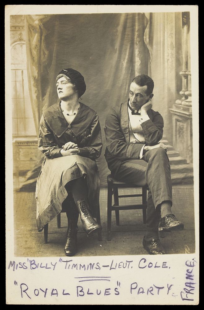 Two soldiers, one in drag called "Miss Billy Timmins", are seated in front of a painted backdrop. Photographic postcard…