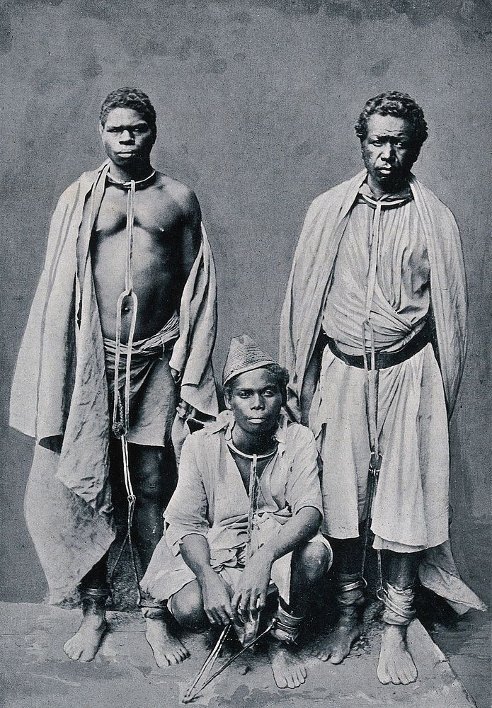 Three men in Madagascar have chains around them that join their necks and ankles. Process print.