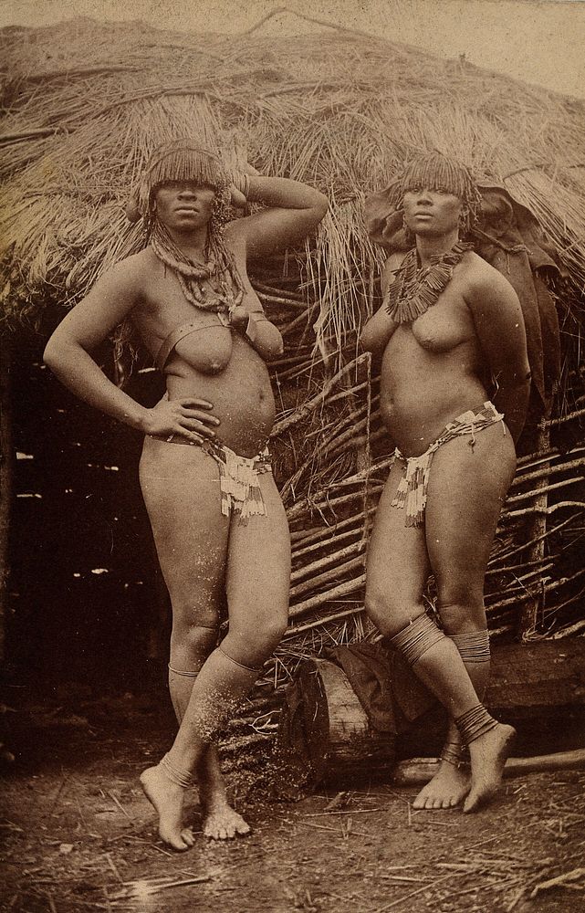 South Africa: Zulu women, bare-chested and wearing ornamental jewellery, posing outside a mud hut. Photograph, 1880/1890.