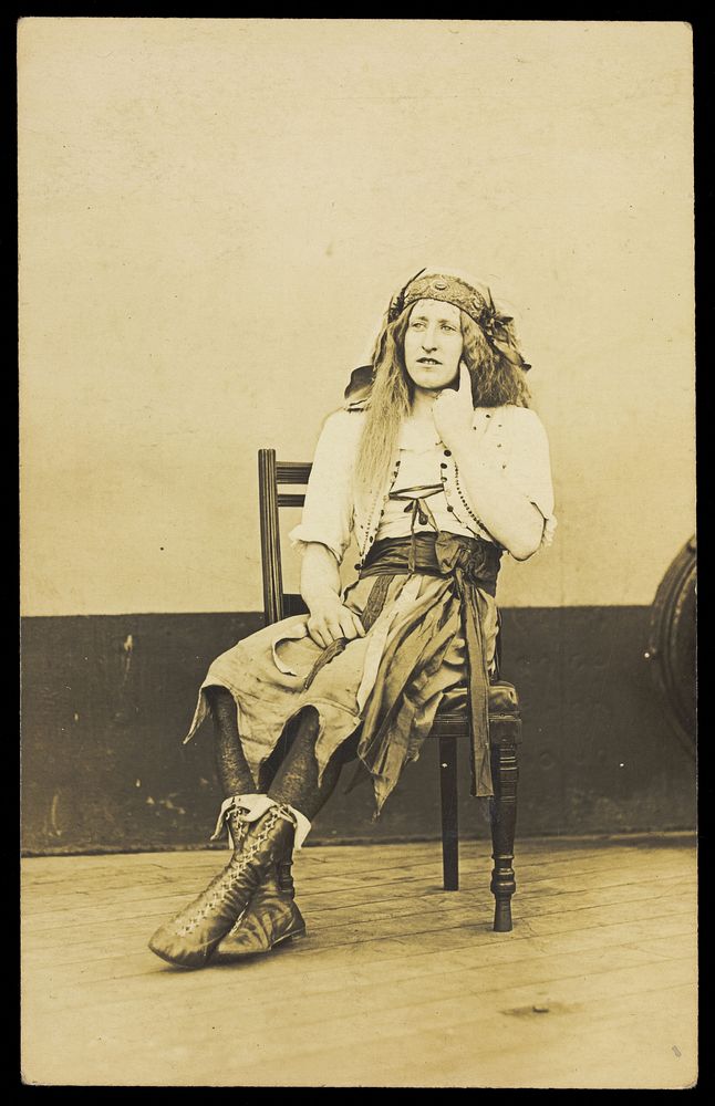 A man in drag sits pensively on a chair, on the deck of a ship. Photographic postcard, 191-.
