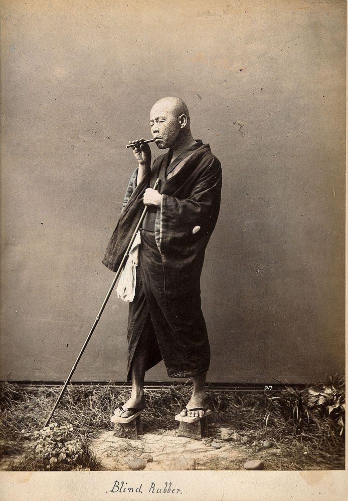 A blind Japanese masseur with a pipe in his mouth and a long pole in his hand. Photograph.