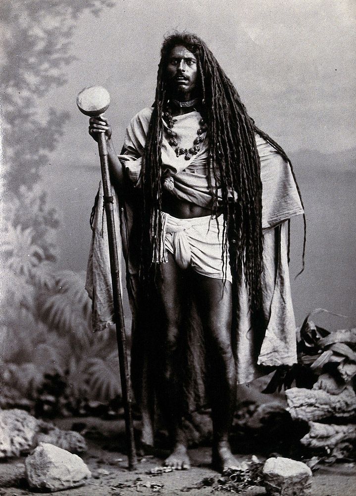 An Indian man with long hair, carrying a staff, in a studio setting. Photograph, ca.1900.