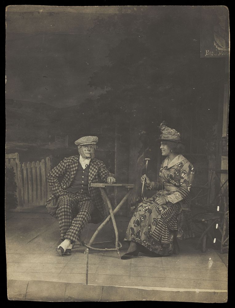 Two sailors, one in drag, sit on stage at a table, in front of very detailed scenery. Photographic postcard, 191-.