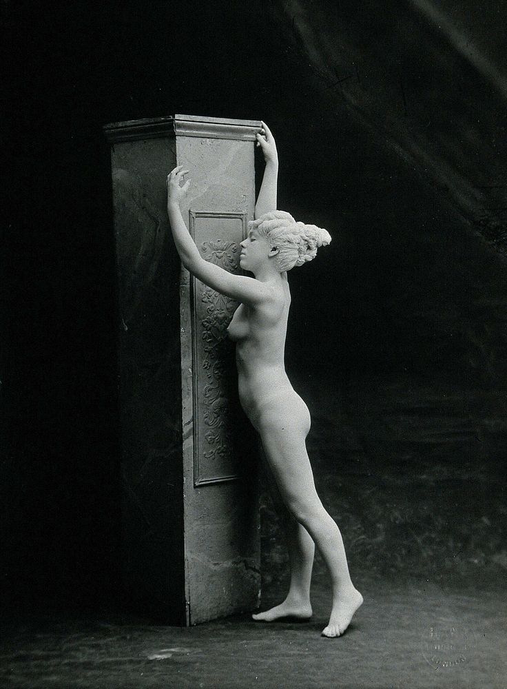 A young woman, posing naked in a classical attitude, against a square column in a photographic studio. Photograph…