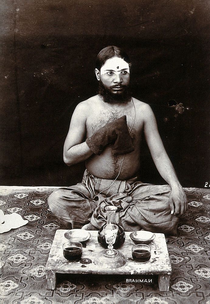 An Indian man wearing a Parsi string and make-up, squatting on a rug with his hand in a rosary bag, in a studio setting.…