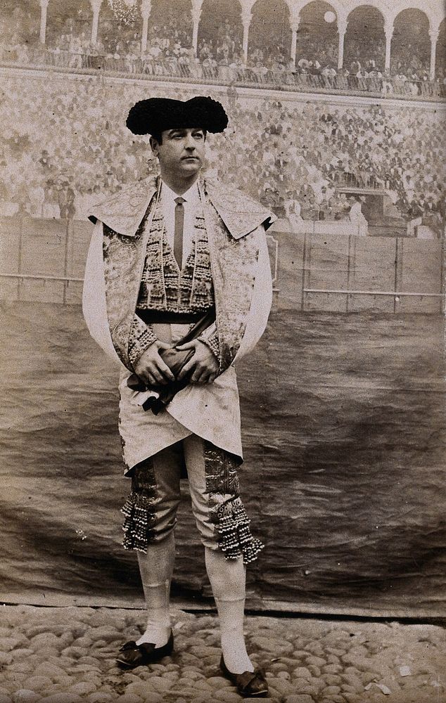 A bullfighter, posing in front of a painted backdrop of a bullring, in a cobbled courtyard. Photograph, first quarter of the…