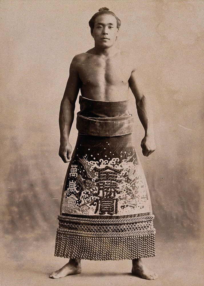 A young sumo wrestler posing in a photographic studio, wearing a richly decorated apron, in front of a plain backdrop.