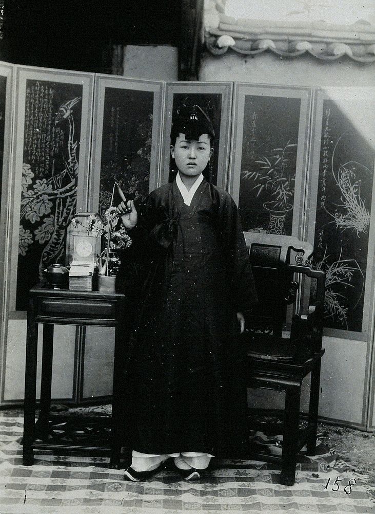 A young woman wearing a plain robe and a tasselled hat, standing in front of a painted screen.