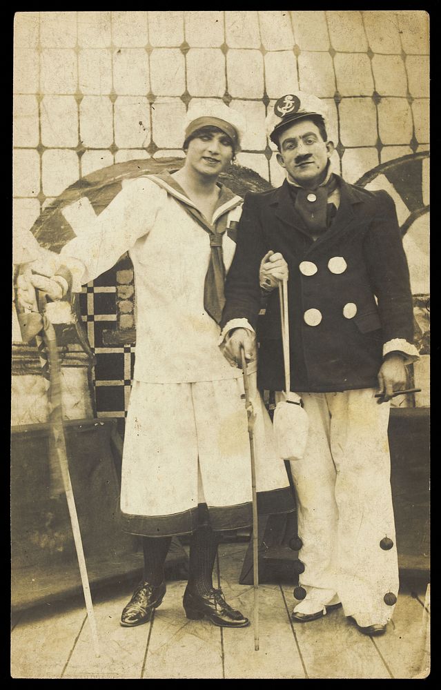 Two sailors, one in drag, pose on deck holding props. Photographic postcard, 191-.