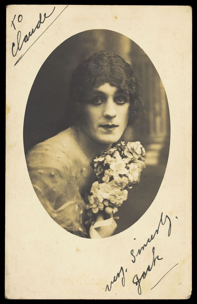 A man in drag wears make-up and is holding flowers, posing for his portrait. Photographic postcard, ca. 1918.