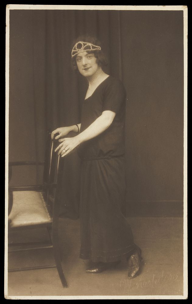 A performer in drag, wearing a black dress and tiara, leaning on a chair. Photographic postcard by W. Aspden, 1924.
