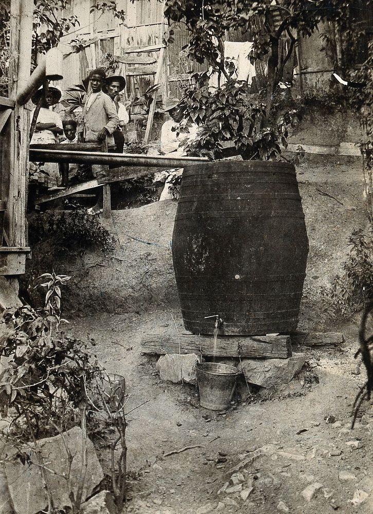 A mosquito-proof water barrel with a tap; residents in the background. Photograph, 1900/1920.