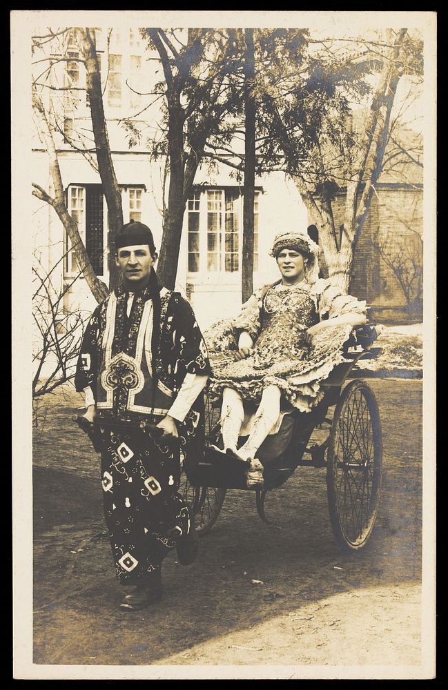 A man in ornate drag sits in a dog-cart and is pulled along the road by a man dressed in Chinese patterned robes and hat.…