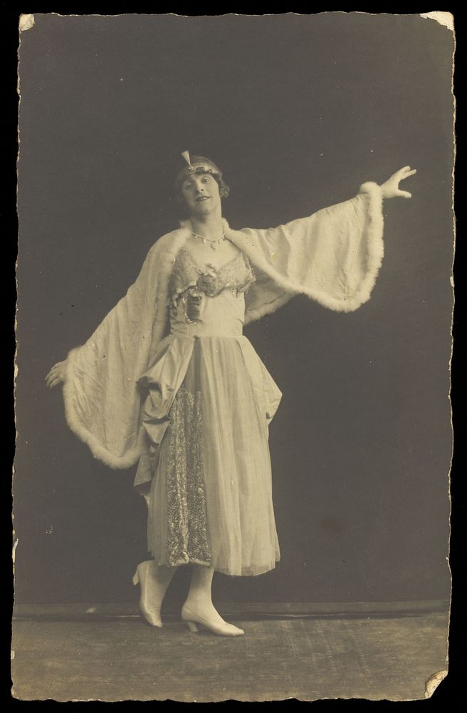 A prisoner of war in drag, wearing a dress and white cloak, poses mid-movement with arms outstretched. Photographic postcard…