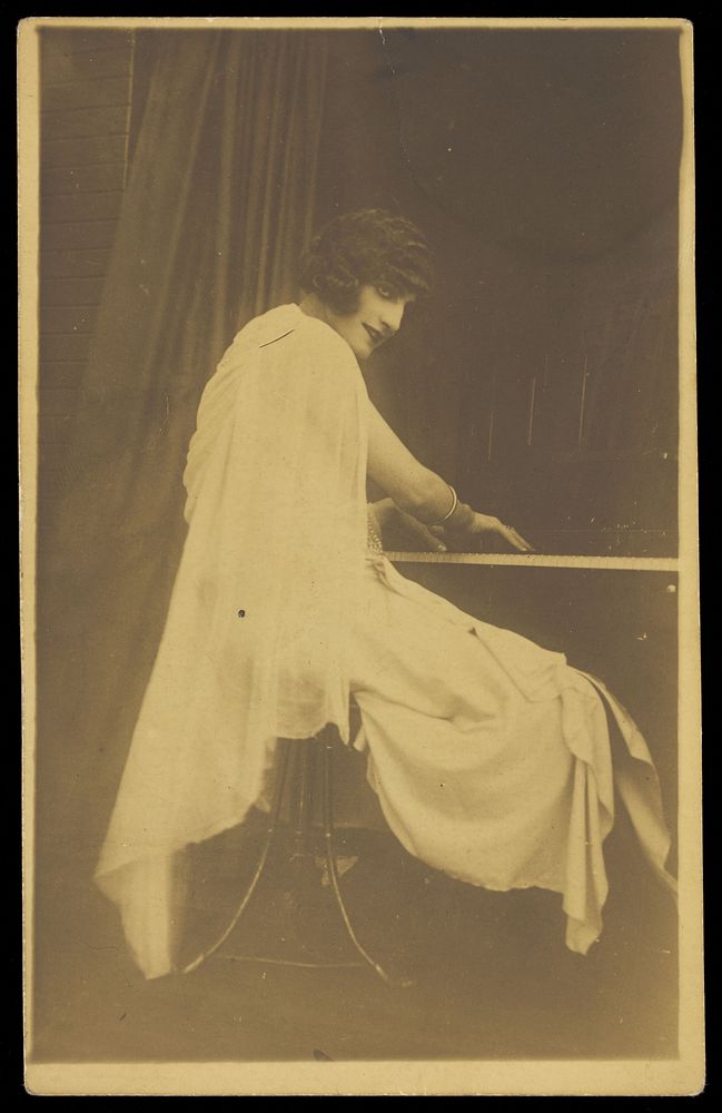 A man wearing a white dress sits playing the piano. Photographic postcard, 192-.