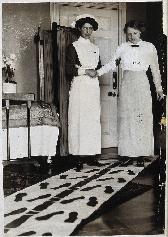 A nurse steadying a woman by holding her right arm, while the woman attempts to walk along a mat marked with footprints.…