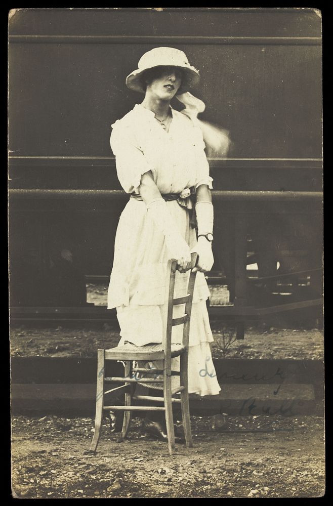 An soldier in a concert party poses in drag, wearing white and holding a chair, standing in front of a train. Photographic…