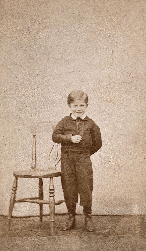 A boy smiling and standing next to a wooden chair. Photograph.