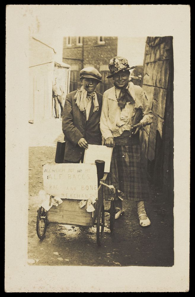 Two young men, one in drag, playing rag and bone merchants. Photographic postcard, 192-.