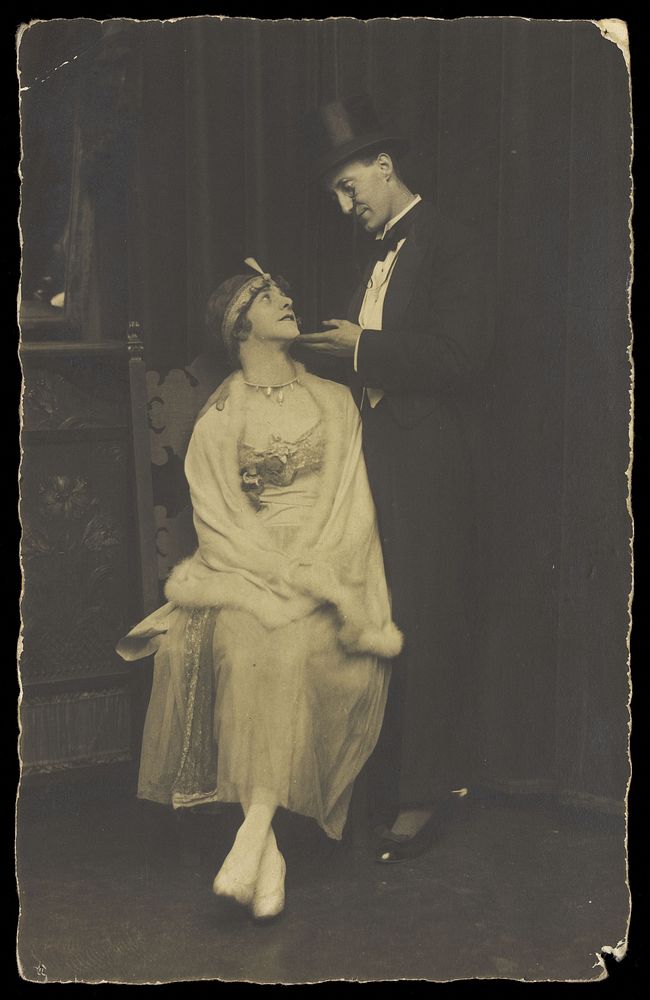 Two actors pose on stage: a man wearing top hat and tails gazes down at a man in drag. Photographic postcard, 192-.