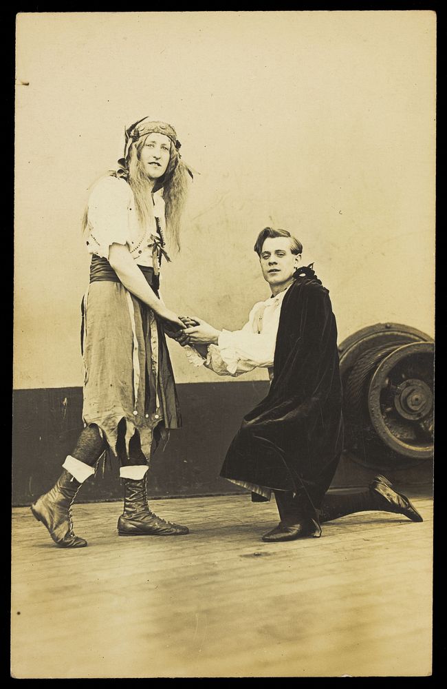 Two amateur actors embrace on the deck of a ship; one is in drag, the other is kneeling on one knee. Photographic postcard…