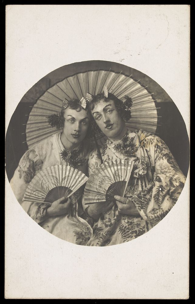 Two young men in drag wearing Japanese costumes, pose together holding fans, underneath a parasol. Photographic postcard…