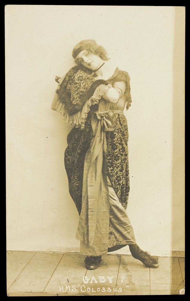 An sailor poses in drag, wearing heavy fabric and a headband. Photographic postcard, ca. 1915-1916.