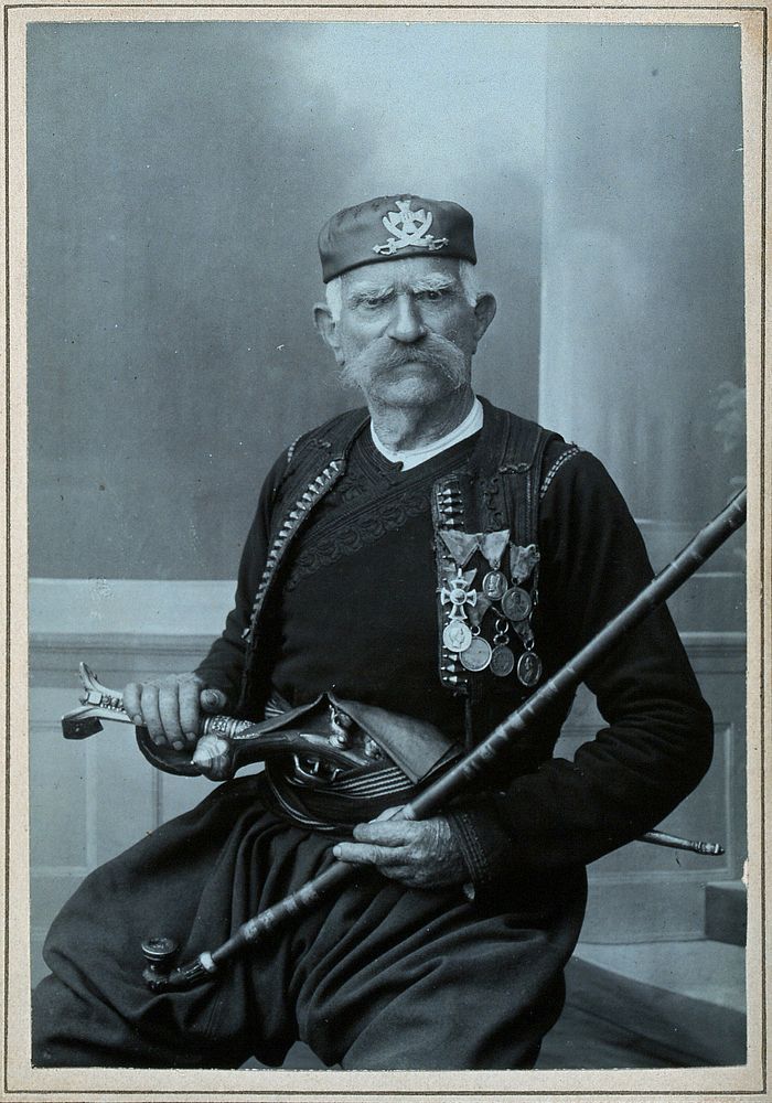 A Montenegrin man wearing national dress with medals, with a pistol in his belt and holding a tobacco pipe.