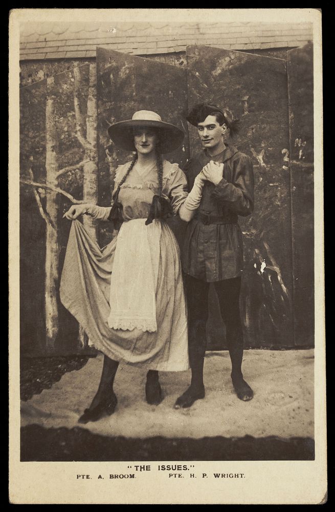 Two members of a military concert party pose on stage: one is in drag with a large hat and pigtails. Photographic postcard…