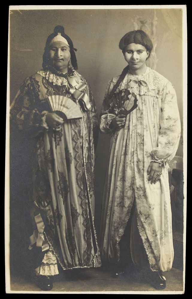 Two men in drag wearing face masks and patterned robes, holding fans. Photographic postcard, ca. 1910.