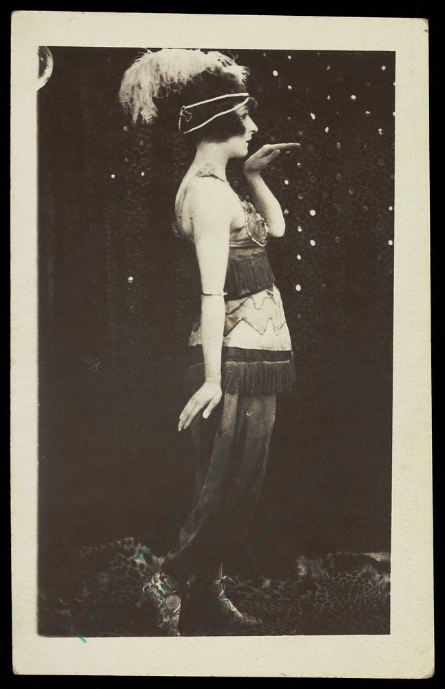 An actor in drag, poses mid-dance, wearing a feathered head garment and a tassled dress. Photographic postcard, 1921.