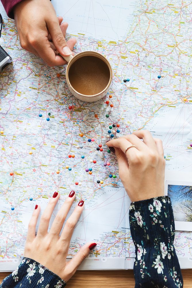 Hands on a map with colorful pins, planning a journey. Map and pins show travel ideas. Coffee cup nearby. Map, pins, and…