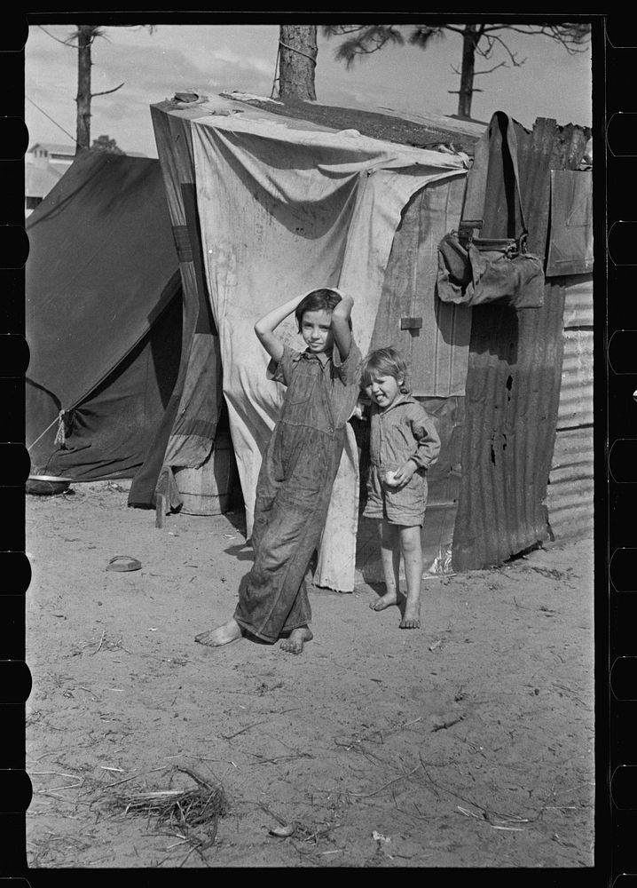 Two children of a migrant fruit worker from Tennessee, standing before their temporary home. This family of eight is camped…