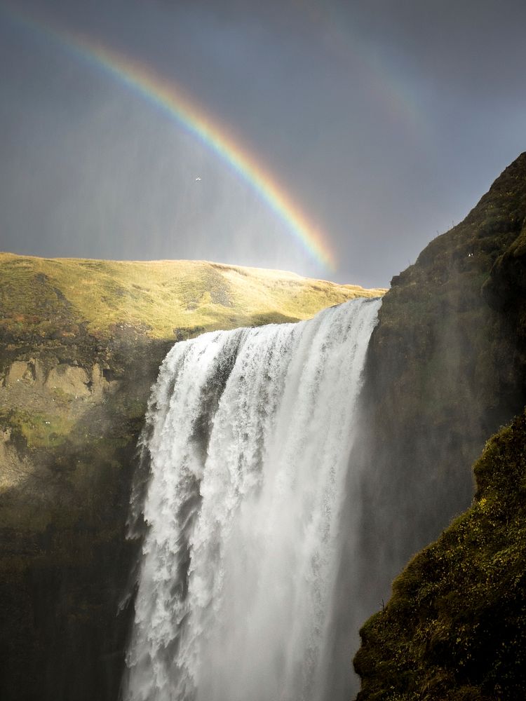 A rainbow over a waterfall pouring down from mossy rocks in Skógafoss. Original public domain image from Wikimedia Commons