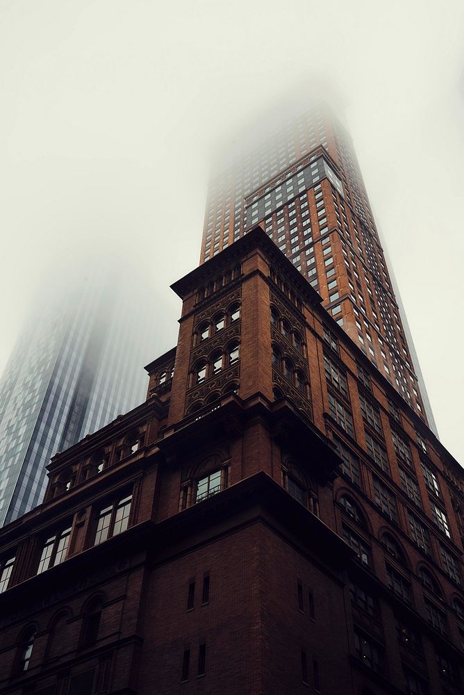 A ground view of a dark brick skyscraper disappearing into fog in New York City. Original public domain image from Wikimedia…