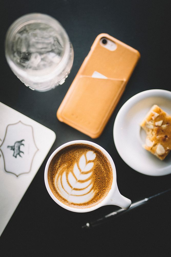 An overhead shot of coffee with latte art on it next to a piece of cake and an iPhone. Original public domain image from…