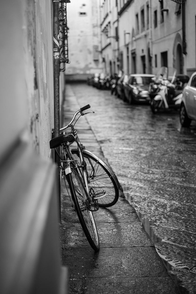 Black and white shot of bike leaning against wall in street with cars and heavy rain. Original public domain image from…