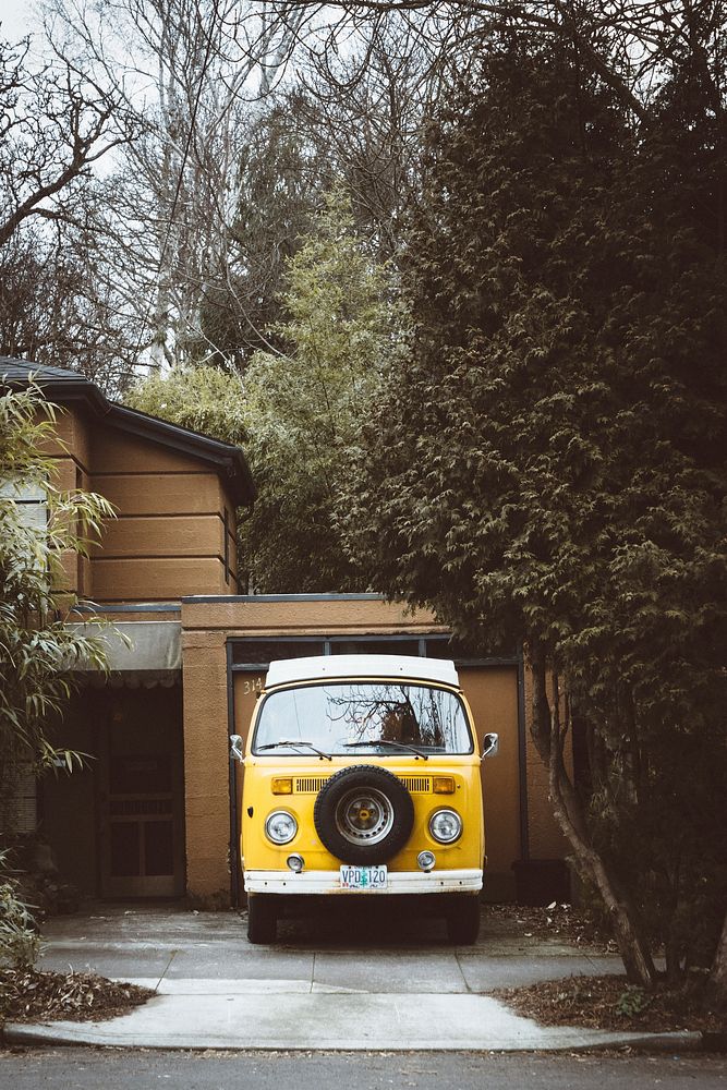 Yellow vintage Volkswagen parked in a driveway of a brown house with trees in the backyard. Original public domain image…