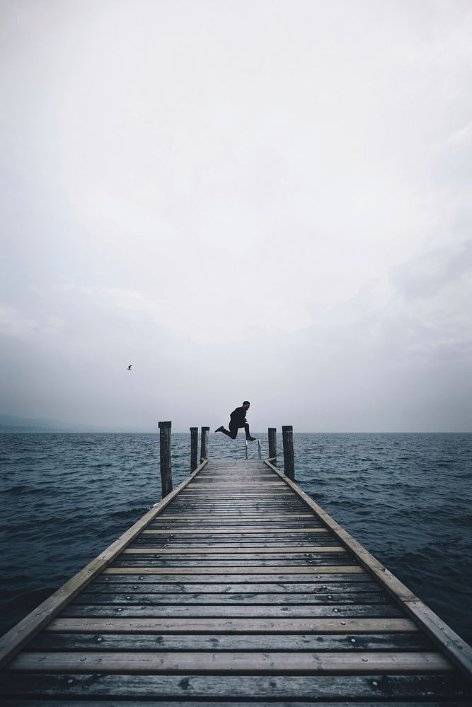 A person jumping in the air on the edge of a dock by a lake in Punta Saint Vigilio on a cloudy day. Original public domain…