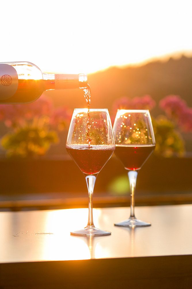 A person pouring red wine into one of two glasses with flowers and the setting sun in the background. Original public domain…