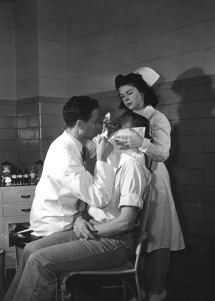 The 1950s historical photograph of an industrial plant physician and nurse examining the eye of an industrial plant worker.
